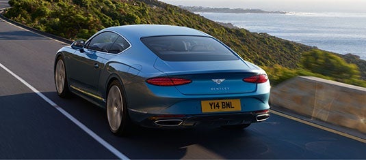 Rear three-quarter view of the Bentley Continental GT Azure driving along a scenic coastal highway, emphasizing its sculpted tail lights, twin exhausts, and sleek fastback design.