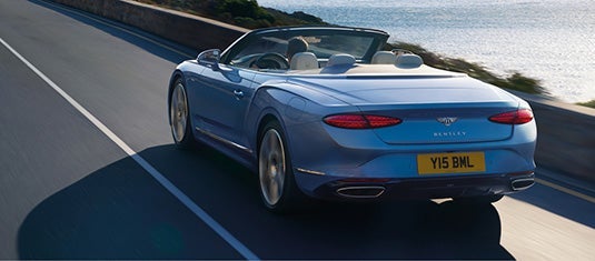 Rear view of the Bentley Continental GTC Azure driving along a seaside highway with the convertible roof down, emphasizing its sculpted rear, twin exhausts, and Bentley badging.