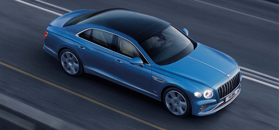 Overhead view of the Bentley Flying Spur Azure driving on an open road, showcasing its elegant blue paintwork, panoramic black roof, and long-wheelbase silhouette.