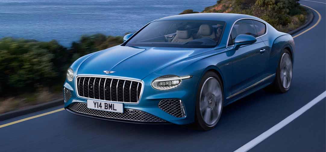 The Bentley Continental GT Azure driving swiftly along a coastal road, its sleek coupe design and wide stance embodying power and dynamic performance.
