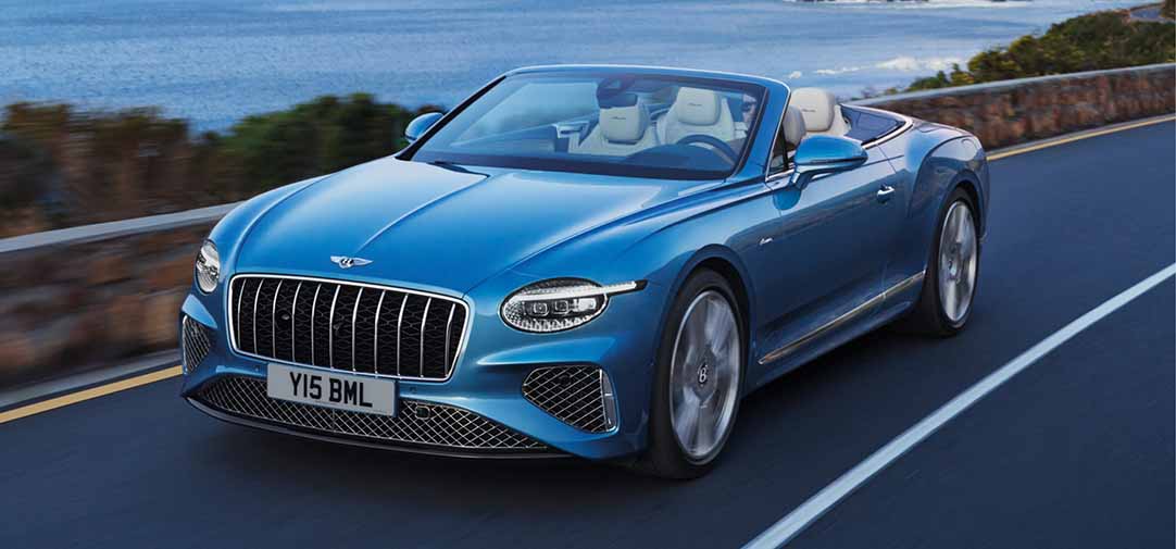 The Bentley Continental GTC Azure convertible in motion with the top down, gliding along a scenic ocean road and capturing the thrill of open-air performance.