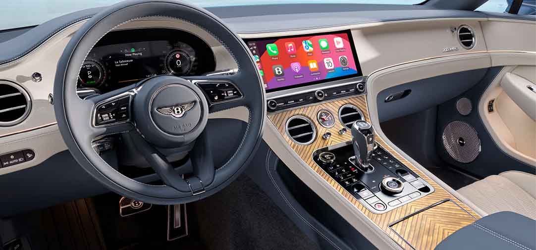 Interior view of the Bentley Continental GT Azure cockpit, showcasing the digital instrument cluster, Apple CarPlay touchscreen, and a wood veneer center console with precision controls.