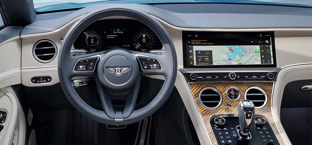 Steering wheel and dashboard view of the Bentley Continental GTC Azure, featuring a full digital instrument cluster, large touchscreen with built-in navigation, and elegant wood veneer detailing