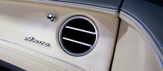 Close-up of the Bentley Continental GTC Azure dashboard, highlighting the Azure emblem beside a chrome-trimmed circular air vent and contrast-stitched leather trim.