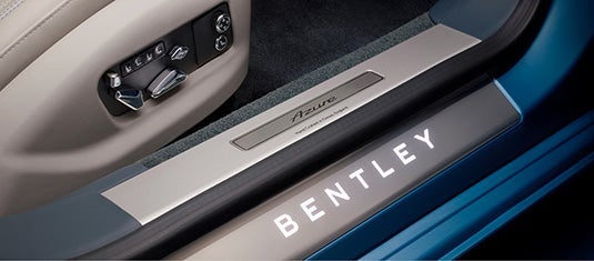 Illuminated Bentley door sill plate with Azure branding next to chrome seat adjustment controls, set against soft-touch leather and carpet trim.