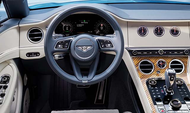 Driver’s view of the Bentley Continental GT cabin, showing the multifunction steering wheel, digital dash, and premium wood veneer dashboard with rotary dials.
