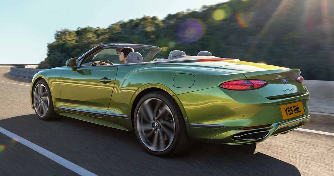 Rear view of a green Bentley Continental GTC convertible driving along a scenic road with the top down and sun flare highlighting the sleek body lines.