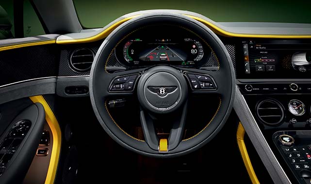 Driver’s cockpit of a Bentley Continental GTC featuring a digital instrument cluster, multifunction steering wheel, and yellow-accented carbon fiber trim.