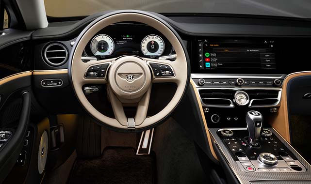 Driver’s seat and dashboard of the Bentley Flying Spur Mulliner, highlighting a beige steering wheel, digital instrument cluster, and touchscreen interface with Apple CarPlay integration.