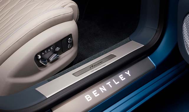 Detail shot of the Bentley Flying Spur Azure door sill illuminated with the “BENTLEY” logo and “Azure” badge beside the beige leather seat and electronic seat controls.