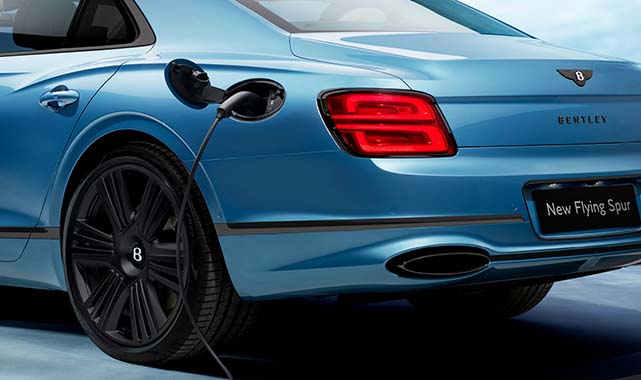 Rear quarter view of a blue Bentley Flying Spur Azure plug-in hybrid connected to a charging cable, with bold black wheels.