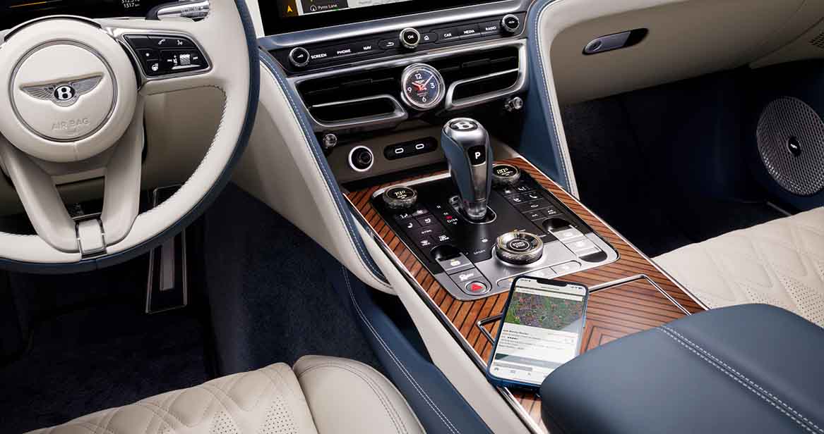 Interior of Bentley Flying Spur featuring cream and blue leather, touchscreen navigation, rotary gear selector, and a smartphone resting on the handcrafted wood veneer console.