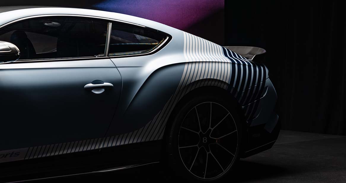 Low-key side profile shot of Bentley Continental GT Supersports in showroom lighting, highlighting sculpted rear fender flares, racing stripe graphics, and large alloy wheels.